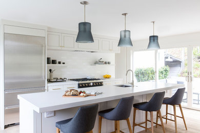 This is an example of a country kitchen in San Francisco with a single-bowl sink, flat-panel cabinets, grey cabinets, quartz benchtops, white splashback, ceramic splashback, black appliances, light hardwood floors, with island, brown floor and grey benchtop.