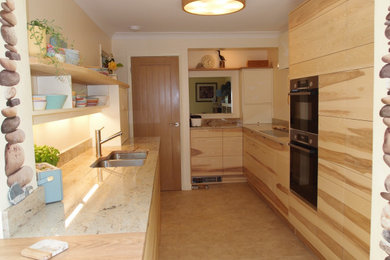 Small modern galley enclosed kitchen in Cornwall with a double-bowl sink, flat-panel cabinets, light wood cabinets, granite worktops, multi-coloured splashback, glass sheet splashback, black appliances, porcelain flooring, a breakfast bar, beige floors, beige worktops and feature lighting.