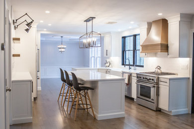 Example of a large transitional medium tone wood floor kitchen design in Boston with a farmhouse sink, recessed-panel cabinets, white cabinets, quartz countertops, white backsplash, subway tile backsplash, stainless steel appliances, an island and white countertops