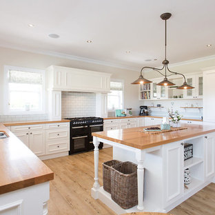 Design ideas for a mid-sized country l-shaped kitchen pantry in Christchurch with a double-bowl sink, louvered cabinets, white cabinets, wood benchtops, white splashback, brick splashback, black appliances, light hardwood floors, with island, brown floor and brown benchtop.