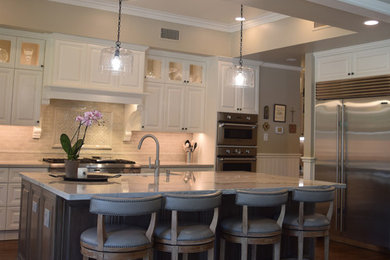 Mid-sized transitional l-shaped dark wood floor and brown floor open concept kitchen photo in Houston with a farmhouse sink, raised-panel cabinets, white cabinets, quartzite countertops, beige backsplash, ceramic backsplash, stainless steel appliances, an island and white countertops