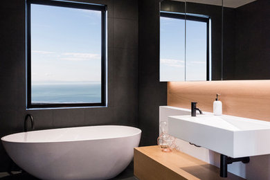 Design ideas for a mid-sized contemporary bathroom in Christchurch with light wood cabinets, a freestanding tub, an open shower, black tile, porcelain tile, porcelain floors, a wall-mount sink, solid surface benchtops, grey floor and an open shower.