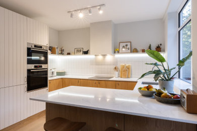 Medium sized contemporary u-shaped kitchen/diner in London with a submerged sink, louvered cabinets, light wood cabinets, quartz worktops, white splashback, ceramic splashback, stainless steel appliances, light hardwood flooring, a breakfast bar, brown floors and white worktops.