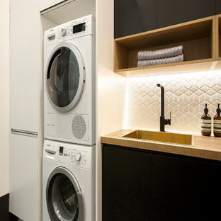 This is an example of a modern single-wall utility room in Melbourne with a built-in sink, flat-panel cabinets, black cabinets, laminate countertops, white walls and a stacked washer and dryer.