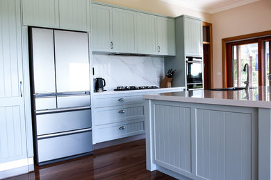 This is an example of a large traditional galley kitchen in Wollongong with a drop-in sink, shaker cabinets, green cabinets, quartz benchtops, white splashback, stone slab splashback, black appliances, medium hardwood floors, a peninsula, brown floor and white benchtop.