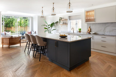 Photo of a large classic galley open plan kitchen in London with a built-in sink, shaker cabinets, beige cabinets, quartz worktops, grey splashback, engineered quartz splashback, black appliances, medium hardwood flooring, an island, beige worktops and feature lighting.