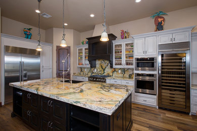 Mid-sized elegant l-shaped bamboo floor and brown floor eat-in kitchen photo in Phoenix with a farmhouse sink, raised-panel cabinets, white cabinets, multicolored backsplash, stone slab backsplash, stainless steel appliances and an island