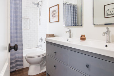 Design ideas for a mid-sized country kids bathroom in Omaha with furniture-like cabinets, grey cabinets, a drop-in tub, a shower/bathtub combo, a two-piece toilet, white tile, ceramic tile, white walls, ceramic floors, an integrated sink, solid surface benchtops, brown floor, a shower curtain and white benchtops.