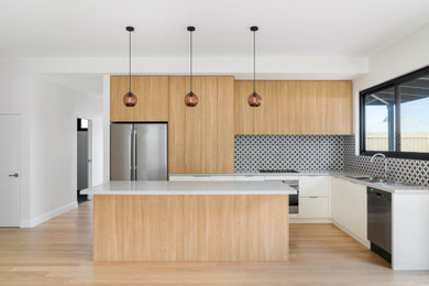 This is an example of a mid-sized modern l-shaped open plan kitchen in Melbourne with a double-bowl sink, shaker cabinets, light wood cabinets, quartz benchtops, black splashback, ceramic splashback, stainless steel appliances, medium hardwood floors, with island, brown floor and white benchtop.