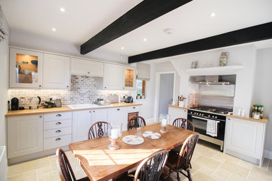This is an example of a mid-sized country l-shaped eat-in kitchen in London with shaker cabinets, grey cabinets, wood benchtops, black appliances, beige floor, an integrated sink, multi-coloured splashback, brick splashback and limestone floors.