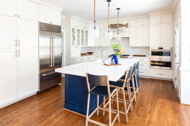 Inspiration for a mid-sized transitional l-shaped medium tone wood floor and brown floor open concept kitchen remodel in Richmond with a farmhouse sink, shaker cabinets, white cabinets, quartz countertops, white backsplash, ceramic backsplash, stainless steel appliances, an island and white countertops