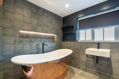 This is an example of a medium sized contemporary family bathroom in Kent with a freestanding bath, a one-piece toilet, grey tiles, porcelain tiles, grey walls, porcelain flooring, a wall-mounted sink, grey floors, a single sink and a freestanding vanity unit.