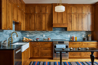 Large elegant l-shaped medium tone wood floor and brown floor eat-in kitchen photo in Minneapolis with a farmhouse sink, shaker cabinets, medium tone wood cabinets, soapstone countertops, blue backsplash, ceramic backsplash, stainless steel appliances, black countertops and an island