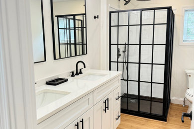 Example of a large farmhouse master white tile laminate floor, brown floor and double-sink corner shower design in Providence with recessed-panel cabinets, white cabinets, a two-piece toilet, white walls, an undermount sink, quartzite countertops, a hinged shower door, white countertops and a built-in vanity