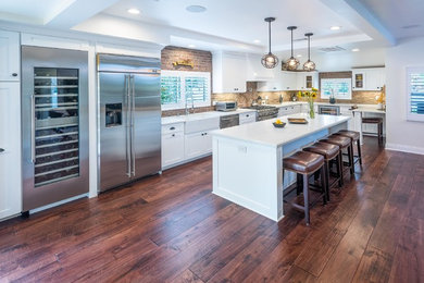 Photo of a mid-sized contemporary l-shaped eat-in kitchen in Los Angeles with a farmhouse sink, shaker cabinets, white cabinets, quartz benchtops, red splashback, brick splashback, stainless steel appliances, dark hardwood floors and with island.