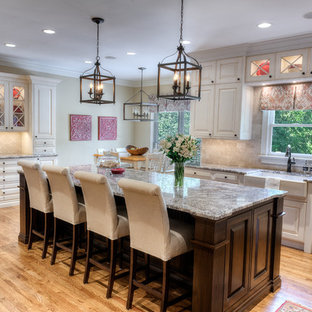 Cette image montre une grande arrière-cuisine traditionnelle en U avec un évier de ferme, un placard avec porte à panneau surélevé, des portes de placard beiges, un plan de travail en granite, une crédence beige, une crédence en carrelage de pierre, un électroménager en acier inoxydable, un sol en bois clair et un îlot central.