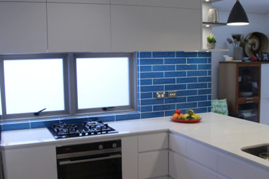 Inspiration for a mid-sized contemporary u-shaped kitchen pantry in Sydney with an undermount sink, recessed-panel cabinets, white cabinets, solid surface benchtops, blue splashback, ceramic splashback, stainless steel appliances, medium hardwood floors, with island, brown floor and white benchtop.