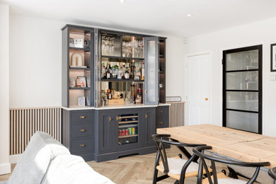 Large modern home bar in Hertfordshire with shaker cabinets, black cabinets, engineered stone countertops, glass sheet splashback, light hardwood flooring, beige floors, white worktops and a feature wall.