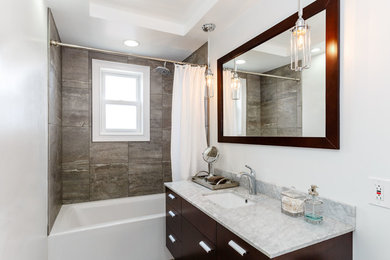 Inspiration for a mid-sized transitional bathroom in New York with an alcove tub, gray tile, porcelain tile, grey walls, porcelain floors, marble benchtops, flat-panel cabinets, dark wood cabinets, a shower/bathtub combo, a one-piece toilet and an integrated sink.