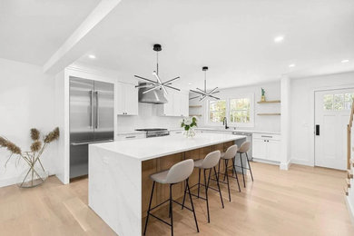 Design ideas for a large l-shaped open plan kitchen in New York with a submerged sink, shaker cabinets, white cabinets, composite countertops, white splashback, metro tiled splashback, stainless steel appliances, light hardwood flooring, an island, beige floors and white worktops.