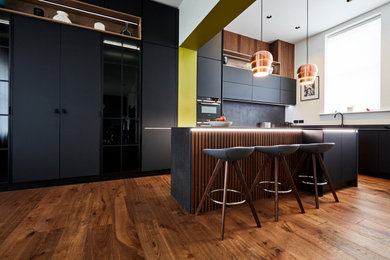 This is an example of a medium sized contemporary kitchen/diner in Other with flat-panel cabinets, black cabinets, black splashback, ceramic splashback, black appliances, medium hardwood flooring, an island, brown floors and black worktops.