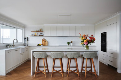 Inspiration for a large transitional open plan kitchen in Sydney with a farmhouse sink, recessed-panel cabinets, white cabinets, quartz benchtops, white splashback, ceramic splashback, black appliances, medium hardwood floors, with island, brown floor and white benchtop.