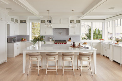 Inspiration for a beach style u-shaped kitchen in Portland Maine with a farmhouse sink, shaker cabinets, white cabinets, grey splashback, subway tile splashback, stainless steel appliances, light hardwood floors, with island, beige floor and white benchtop.