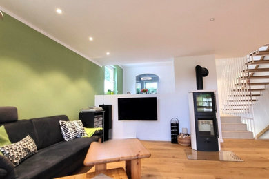 Mittelgroßes, Offenes Modernes Wohnzimmer mit grüner Wandfarbe, braunem Holzboden, Kaminofen, verputzter Kaminumrandung, TV-Wand und eingelassener Decke