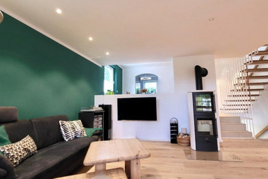 Mittelgroßes, Offenes Modernes Wohnzimmer mit grüner Wandfarbe, braunem Holzboden, Kaminofen, verputzter Kaminumrandung, TV-Wand und eingelassener Decke
