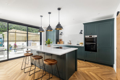 Photo of a large classic single-wall open plan kitchen in Dorset with a built-in sink, shaker cabinets, green cabinets, quartz worktops, white splashback, black appliances, medium hardwood flooring, an island, white worktops, marble splashback, brown floors and feature lighting.