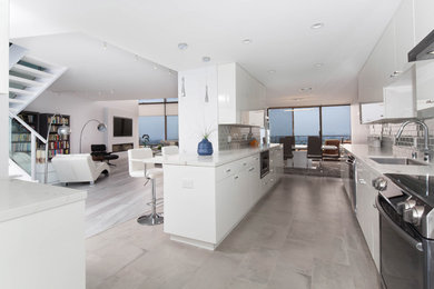 Large trendy galley porcelain tile and gray floor eat-in kitchen photo in Los Angeles with a single-bowl sink, flat-panel cabinets, white cabinets, quartz countertops, gray backsplash, glass tile backsplash, stainless steel appliances, a peninsula and multicolored countertops