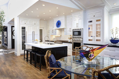 Eat-in kitchen - large contemporary l-shaped medium tone wood floor and brown floor eat-in kitchen idea in Orange County with an undermount sink, recessed-panel cabinets, white cabinets, white backsplash, stainless steel appliances and an island