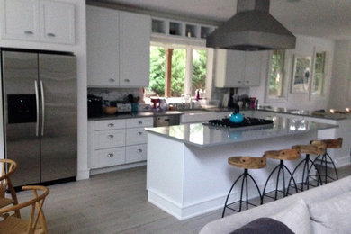 Inspiration for a mid-sized coastal l-shaped light wood floor open concept kitchen remodel in New York with a farmhouse sink, shaker cabinets, white cabinets, solid surface countertops, gray backsplash, stainless steel appliances and an island