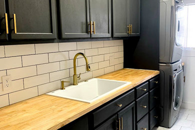 Example of a large classic single-wall ceramic tile and white floor dedicated laundry room design in Louisville with a drop-in sink, shaker cabinets, black cabinets, wood countertops, white backsplash, subway tile backsplash, white walls, a stacked washer/dryer and brown countertops