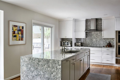 Modelo de cocina de tamaño medio con puertas de armario blancas, salpicadero verde, electrodomésticos de acero inoxidable, suelo de madera en tonos medios, una isla y encimeras grises