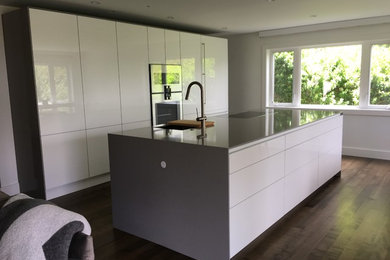 Design ideas for a mid-sized contemporary galley eat-in kitchen in Hawaii with a single-bowl sink, flat-panel cabinets, white cabinets, quartz benchtops, stainless steel appliances, medium hardwood floors, with island, brown floor and grey benchtop.