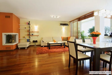 Cette photo montre une grande salle à manger ouverte sur le salon chic avec un mur orange, un sol en bois brun, une cheminée d'angle et un manteau de cheminée en béton.