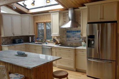 Example of a mid-sized beach style l-shaped light wood floor and brown floor open concept kitchen design in New York with an undermount sink, recessed-panel cabinets, beige cabinets, granite countertops, beige backsplash, stone tile backsplash, stainless steel appliances and an island