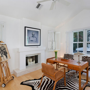 Modelo de despacho actual, de tamaño medio, con paredes blancas, suelo de madera clara, chimenea tradicional, marco de chimenea de yeso, escritorio independiente y suelo beige