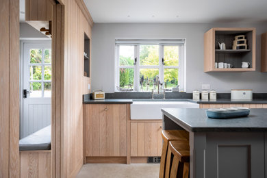 This is an example of a mid-sized beach style galley separate kitchen in Gloucestershire with a farmhouse sink, medium wood cabinets, with island, beige floor and grey benchtop.