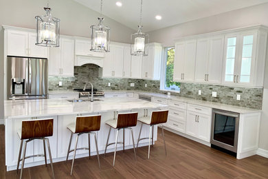 Example of a large trendy l-shaped porcelain tile, brown floor and vaulted ceiling eat-in kitchen design with an undermount sink, shaker cabinets, white cabinets, quartz countertops, gray backsplash, glass tile backsplash, stainless steel appliances, an island and white countertops