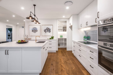 This is an example of a mid-sized traditional galley kitchen pantry in Brisbane with an undermount sink, shaker cabinets, white cabinets, quartz benchtops, window splashback, stainless steel appliances, medium hardwood floors, with island, brown floor and grey benchtop.