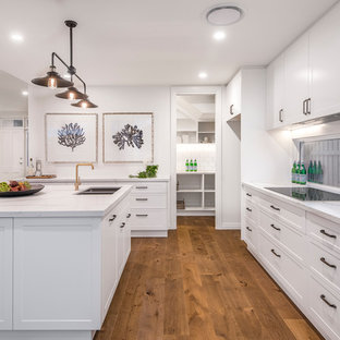 Cette photo montre une arrière-cuisine parallèle chic de taille moyenne avec un évier encastré, un placard à porte shaker, des portes de placard blanches, un plan de travail en quartz modifié, une crédence en fenêtre, un électroménager en acier inoxydable, un sol en bois brun, un îlot central, un sol marron et un plan de travail gris.