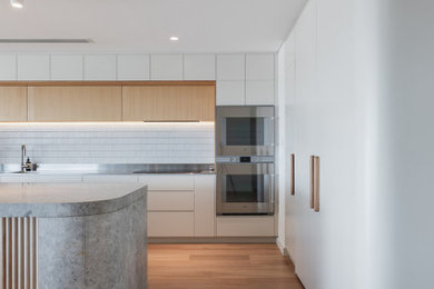 Design ideas for an expansive contemporary l-shaped open plan kitchen in Perth with an integrated sink, white cabinets, marble benchtops, white splashback, ceramic splashback, stainless steel appliances, light hardwood floors, with island and grey benchtop.