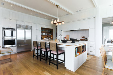 Example of a trendy l-shaped medium tone wood floor and brown floor kitchen design in Other with flat-panel cabinets, white cabinets, white backsplash, an island and white countertops