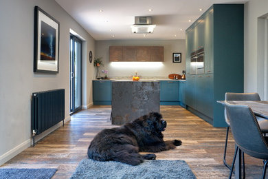 Medium sized contemporary l-shaped open plan kitchen in Other with a built-in sink, flat-panel cabinets, green cabinets, tile countertops, grey splashback, wood splashback, stainless steel appliances, laminate floors, an island, brown floors and brown worktops.