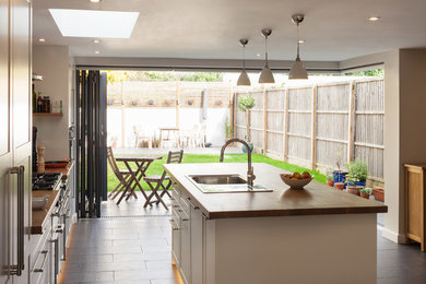 This is an example of a medium sized contemporary galley open plan kitchen in London with a single-bowl sink, recessed-panel cabinets, white cabinets, wood worktops, white splashback, ceramic splashback, stainless steel appliances, ceramic flooring and an island.