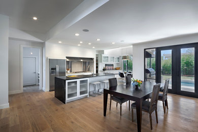 Inspiration for a mid-sized contemporary single-wall medium tone wood floor and brown floor eat-in kitchen remodel in Austin with a farmhouse sink, glass-front cabinets, white cabinets, laminate countertops, metallic backsplash, stainless steel appliances and an island
