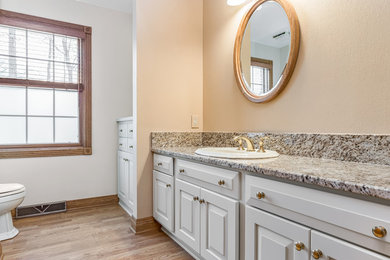 Inspiration for a mid-sized traditional 3/4 bathroom in Milwaukee with raised-panel cabinets, white cabinets, a two-piece toilet, beige walls, medium hardwood floors, a drop-in sink, granite benchtops, brown floor and multi-coloured benchtops.