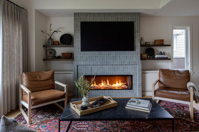 バンクーバーにあるトランジショナルスタイルのおしゃれなリビング (白い壁、無垢フローリング、横長型暖炉、タイルの暖炉まわり、壁掛け型テレビ、茶色い床) の写真